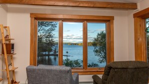 Habitación con un ventanal grande que ofrece vistas a un lago y árboles. Hay dos sillas y una escalera de madera apoyada en la pared.