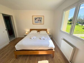A bedroom with a wooden bed, white linens, two bedside tables with lamps, a framed picture on the wall, and a window with a view of greenery.