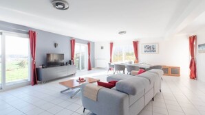 A modern living room with a grey sofa, a coffee table, and a TV. There is a dining area with a table and chairs. The room has large windows with red curtains. - Penn Ar Roz - Large house Pwd access near the sea (Saint-Pabu)