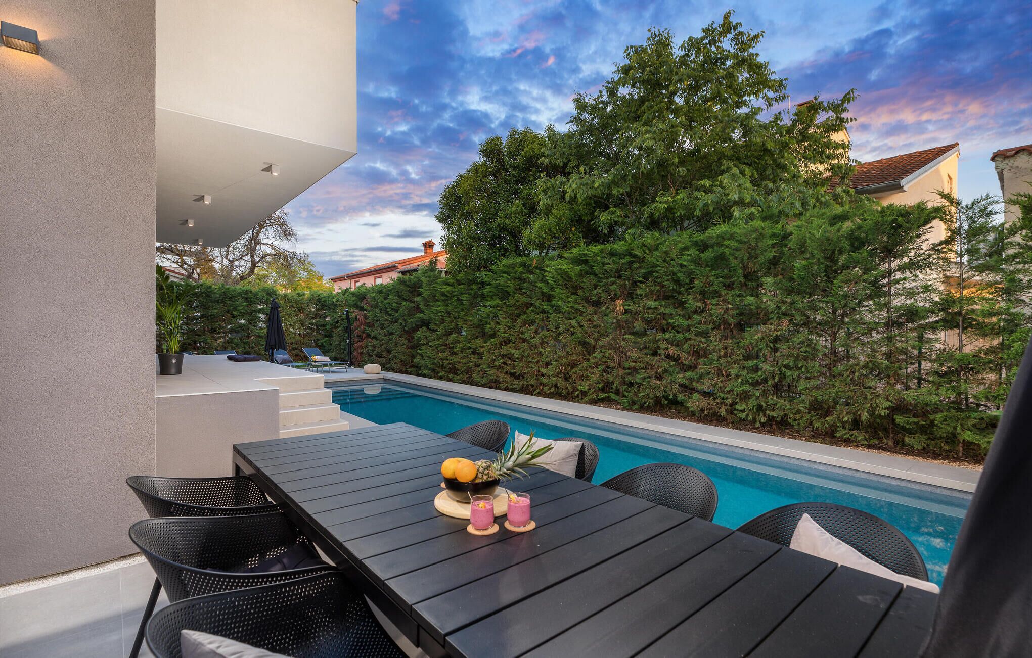 An outdoor pool area with a table and chairs, surrounded by greenery and a view of a building in the background.