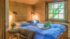 Una habitación de cabaña acogedora con interior de madera, una cama con ropa de cama azul, dos lámparas fijadas a la pared y una ventana con vistas a la vegetación.