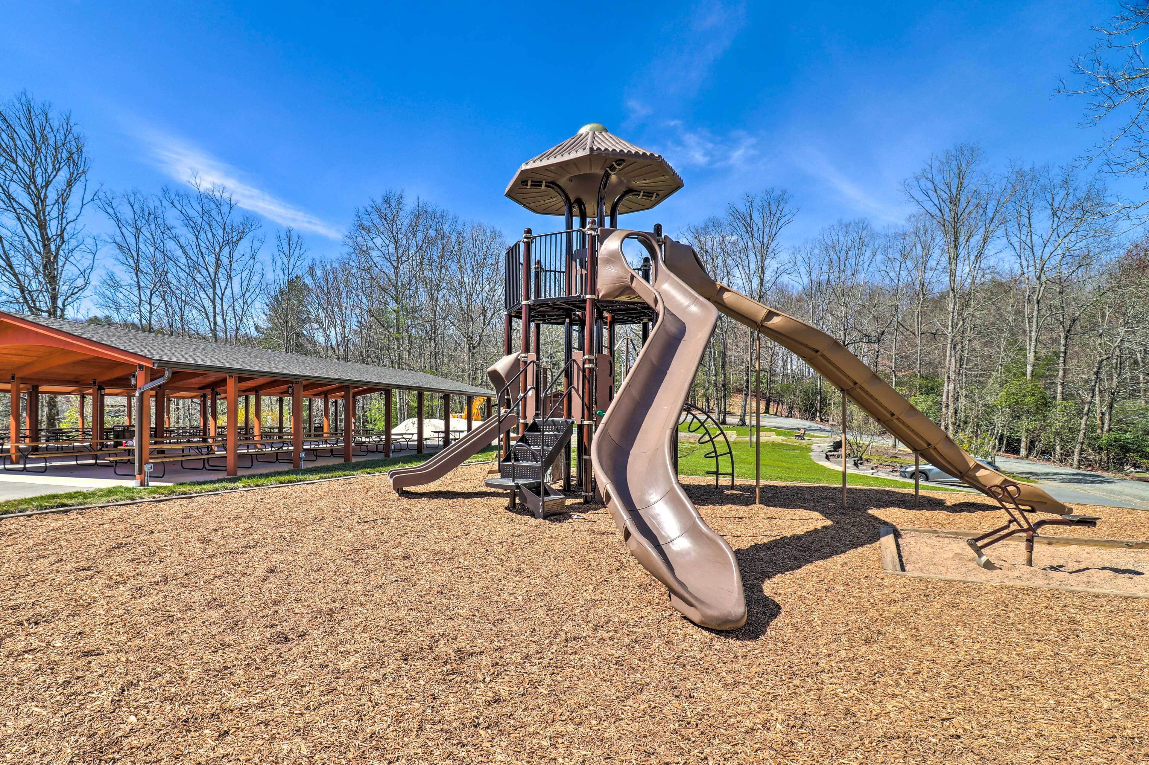 Un parque infantil con tobogán y estructura de escalada, rodeado de árboles y una zona de estar cubierta.