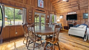 Ein Holzblockhaus-Interieur mit Esstisch, Stühlen, einer Couch, einem Fernseher und einem Kamin.
