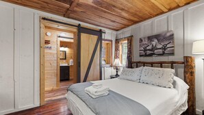Ein Schlafzimmer mit Holzboden, einem Bett mit weißer Bettwäsche, einem Bild an der Wand und einer Tür zu einem Badezimmer.