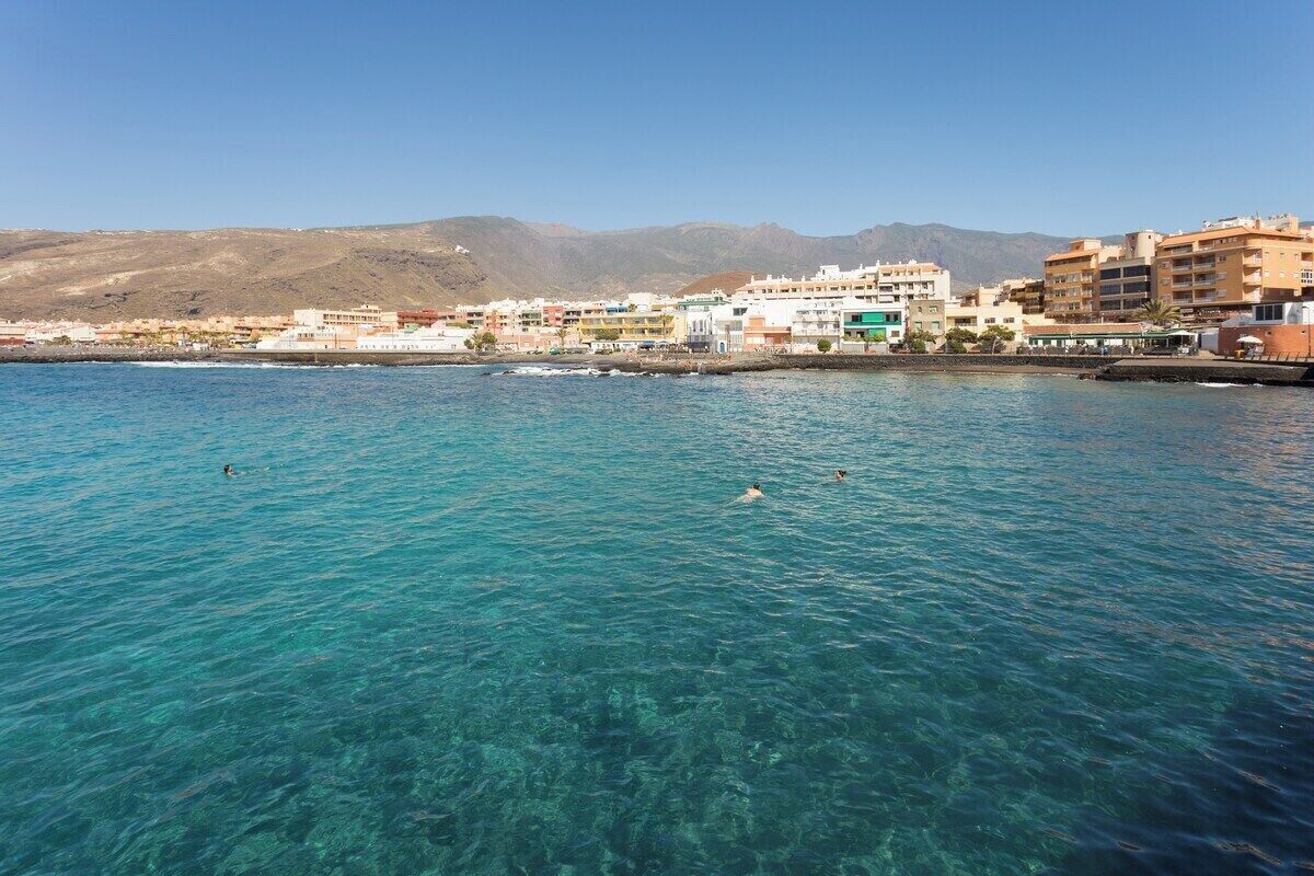 A coastal town with clear blue waters, buildings along the shore, and mountains in the background.