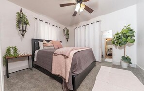 A bedroom with a bed, a ceiling fan, a nightstand with a plant, and a door leading to another room.