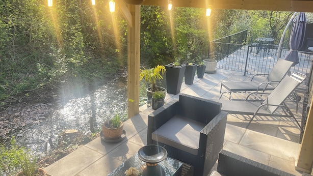 A covered patio with outdoor furniture, potted plants, and a view of a natural area with a stream. - Riverside Two Bedroom Townhouse in Abergavenny with Car Charger. (Abergavenny)