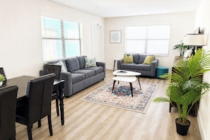 A living room with a grey sofa, a dining table with chairs, a coffee table, and a potted plant.