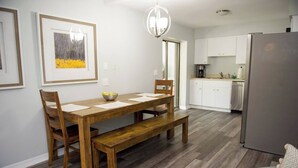 A dining area with a wooden table, benches, and chairs, a kitchen with white cabinets and appliances, and a gray sofa.