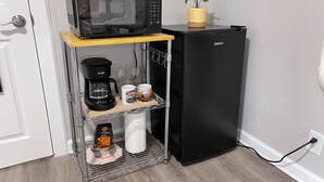 A compact kitchen with a black refrigerator, a microwave on a shelf, a coffee maker, and a small potted plant.