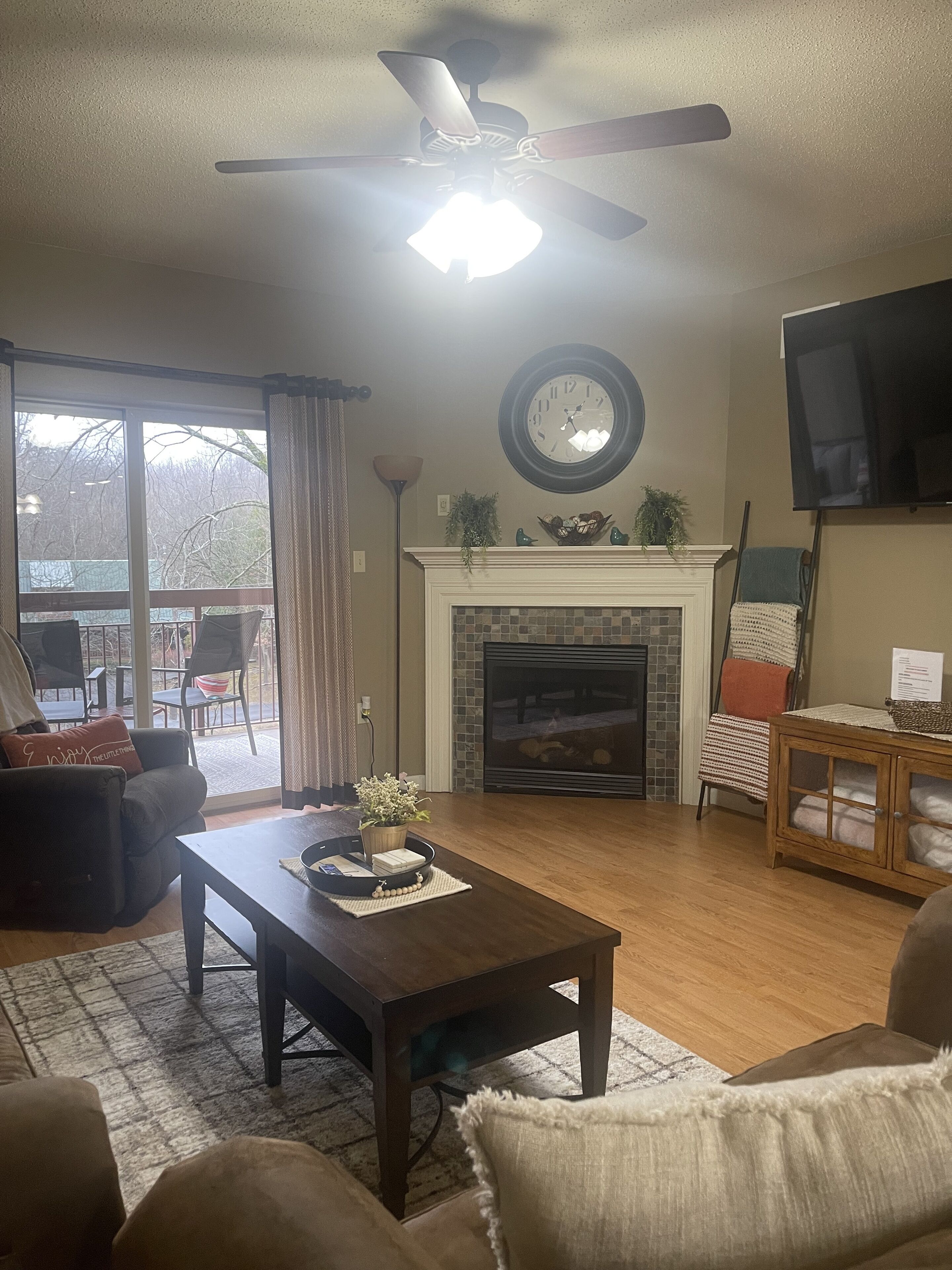 A living room with a fireplace, a ceiling fan, and a television mounted on the wall.