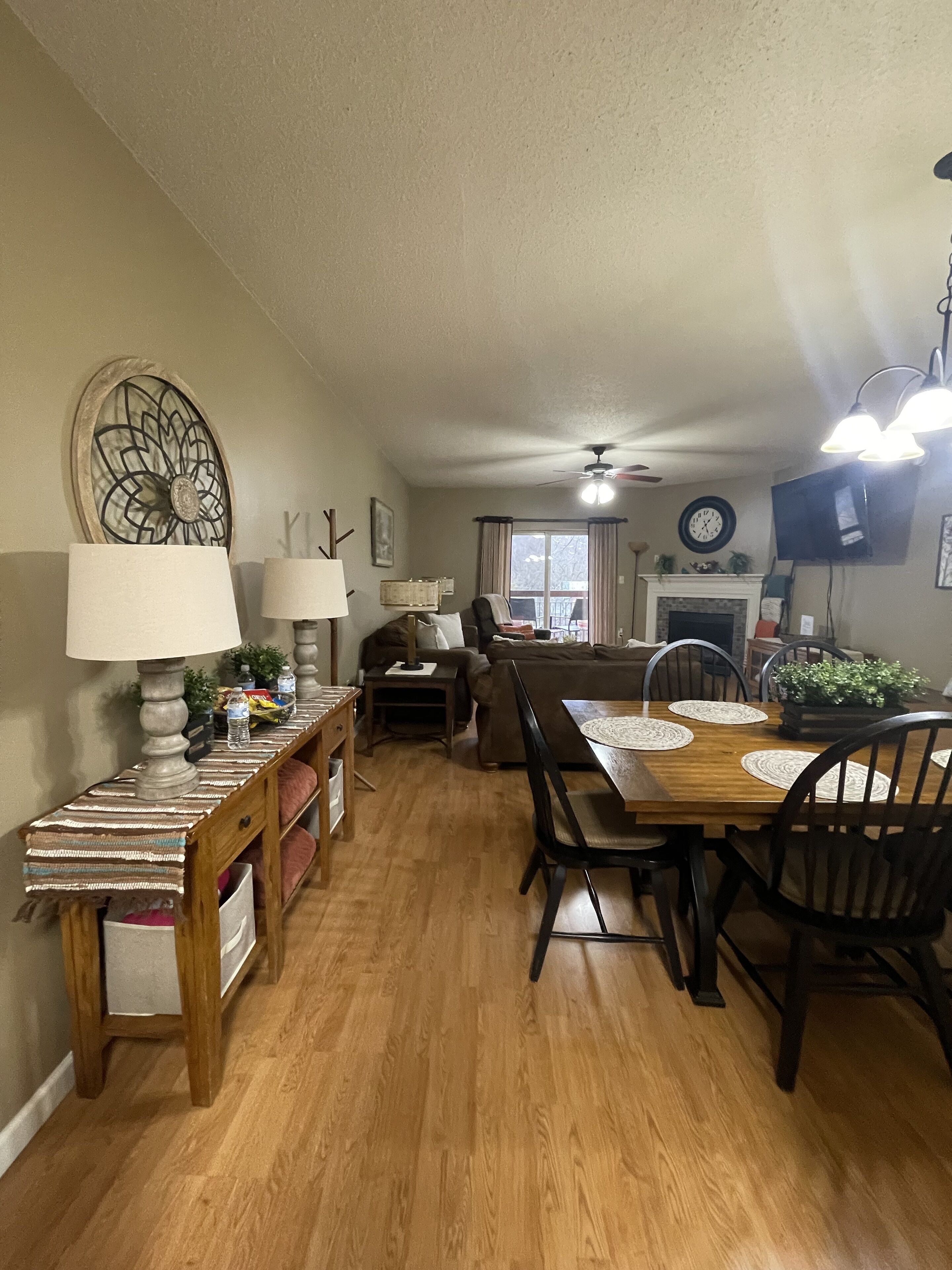 A dining area with a wooden table, chairs, and a chandelier. There is a fireplace and a television mounted on the wall.
