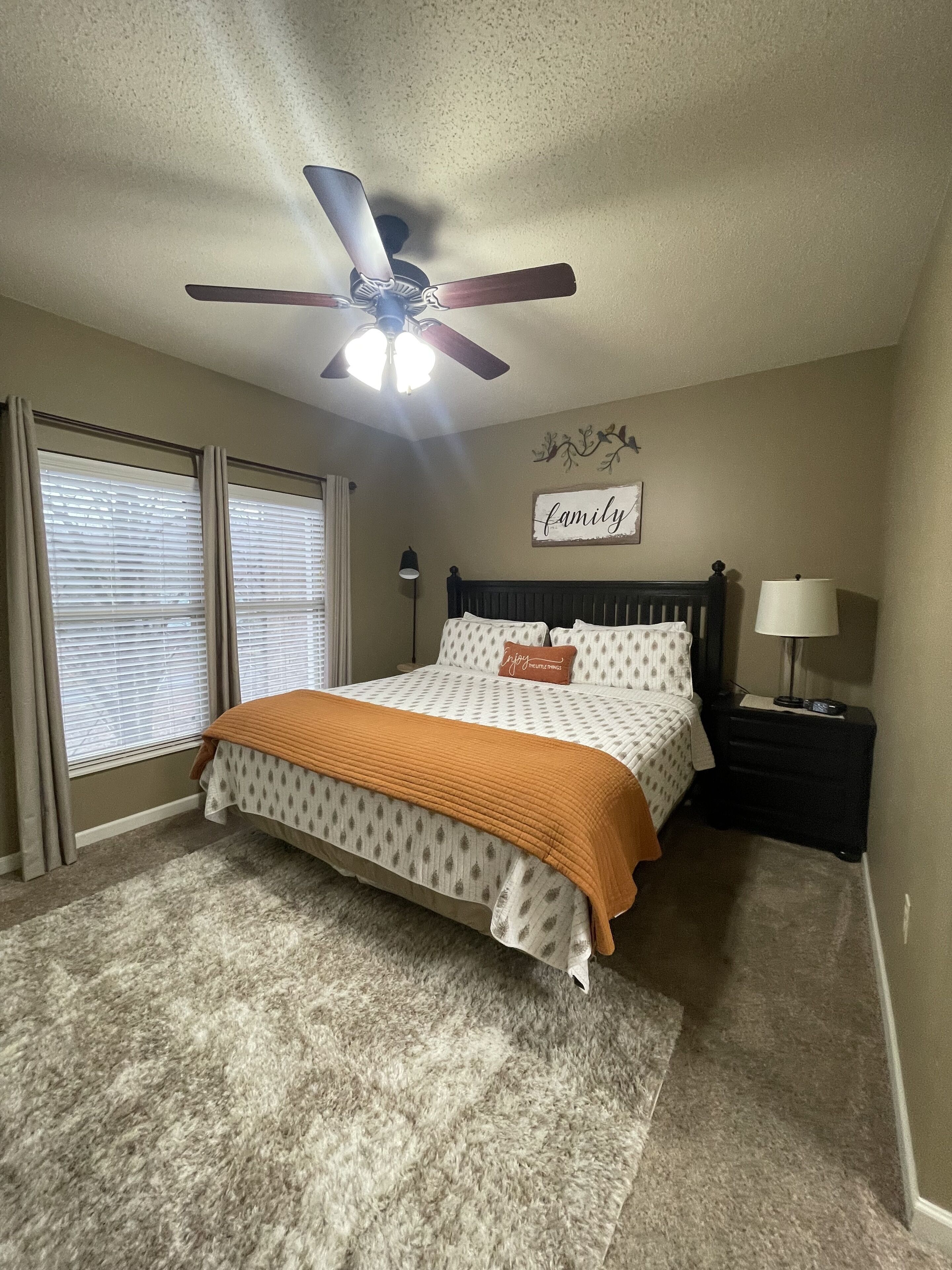 A bedroom with a bed, a ceiling fan, a nightstand, a lamp, and a window with blinds.