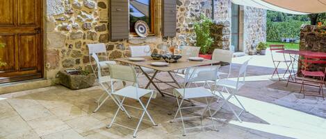 A stone building with a wooden door, a dining area with white chairs and a table, and a covered outdoor seating area.