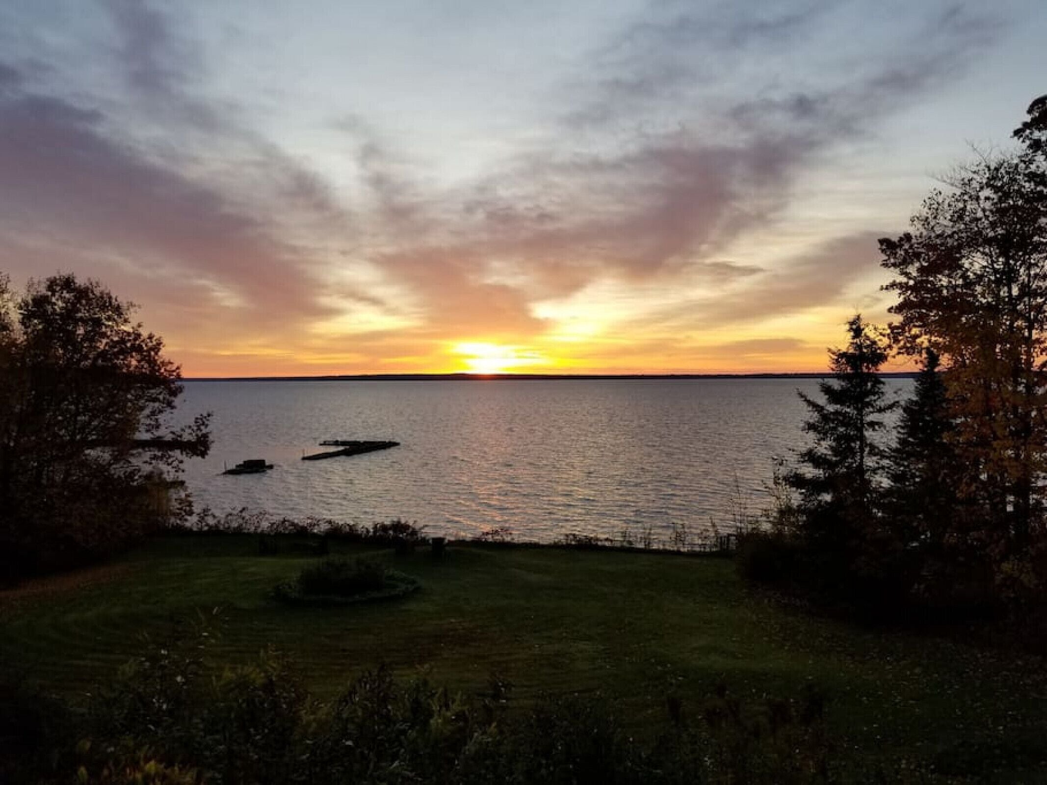 Un coucher de soleil sur une étendue d’eau calme, avec un premier plan composé d’herbe et d’arbres.