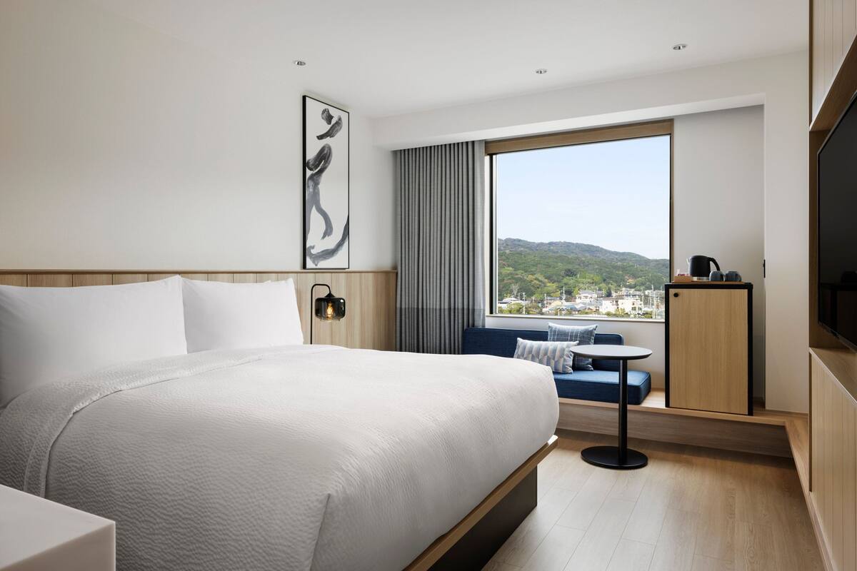 A modern hotel room with a large bed, a small table, a sofa, a TV, and a window with a city view.