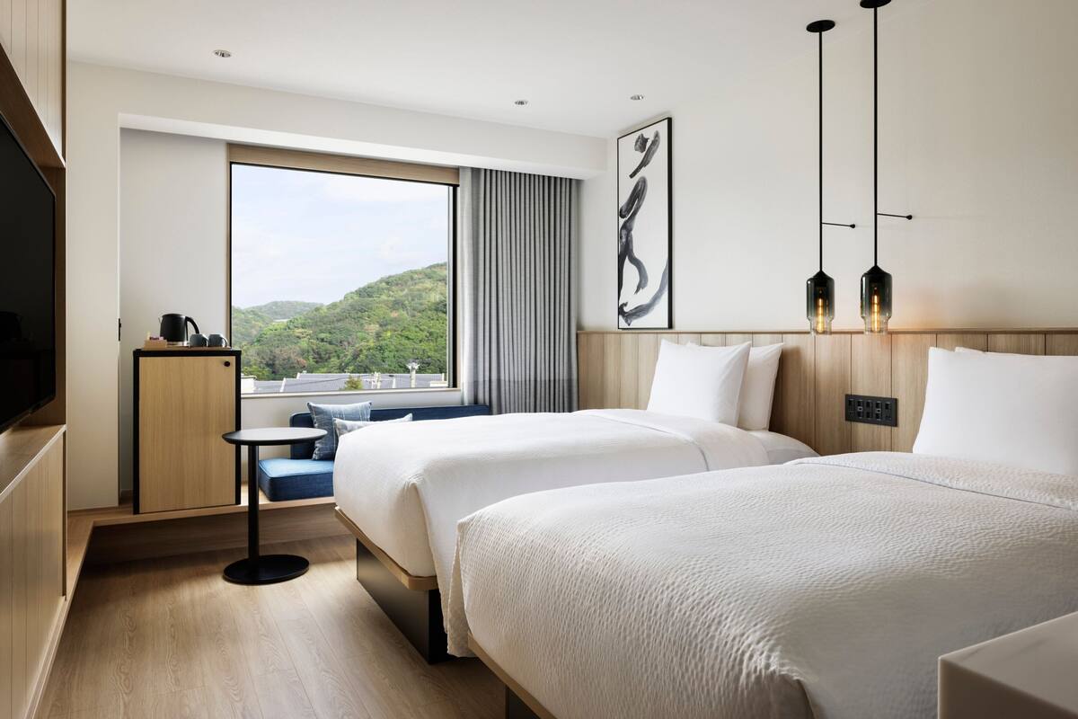 A hotel room with two beds, a small table, a TV, a window with a view of greenery, and a painting on the wall.