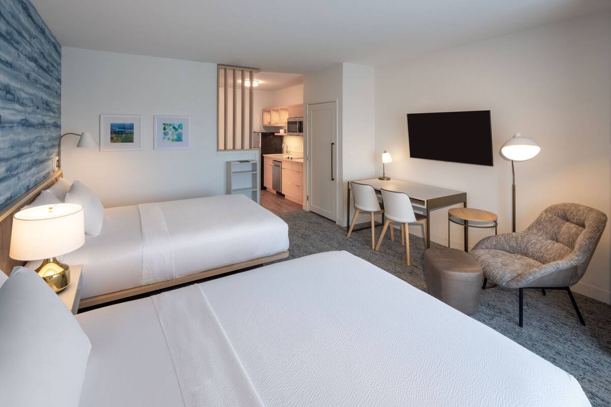 Habitación de hotel con dos camas, un escritorio, una silla, una lámpara y una cocina visible a través de una puerta abierta.