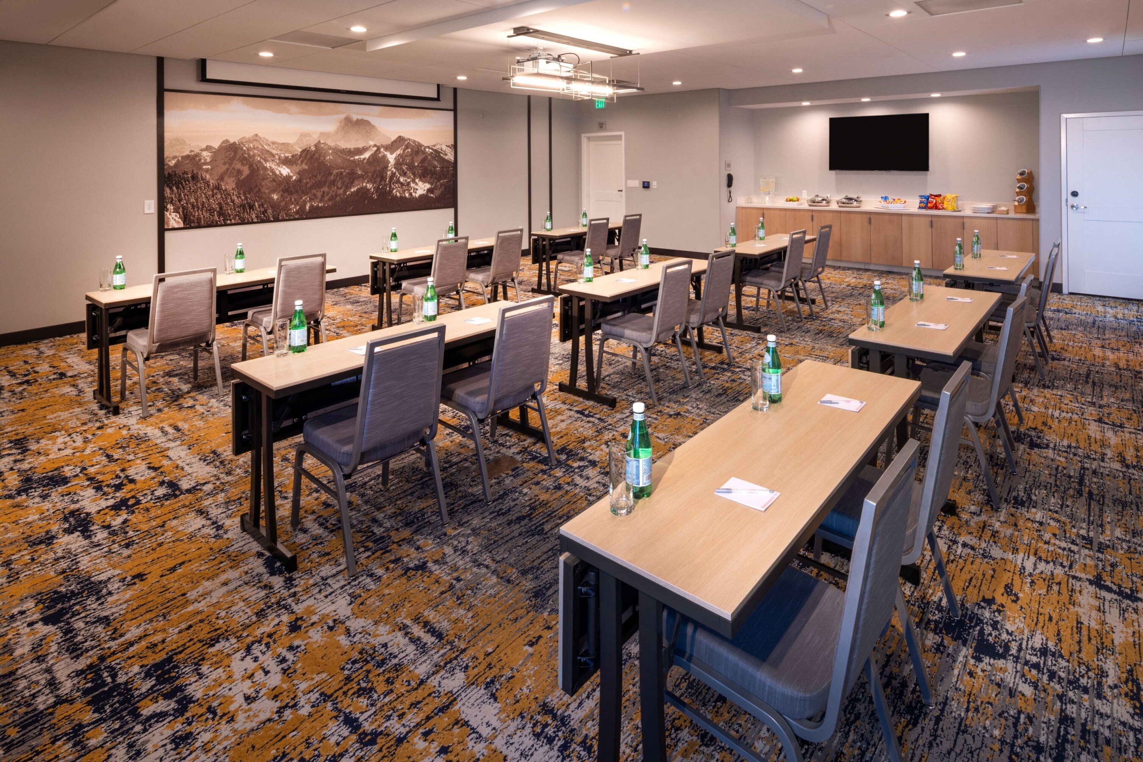 A conference room with multiple tables and chairs, equipped with water bottles, a projector, and a large screen.
