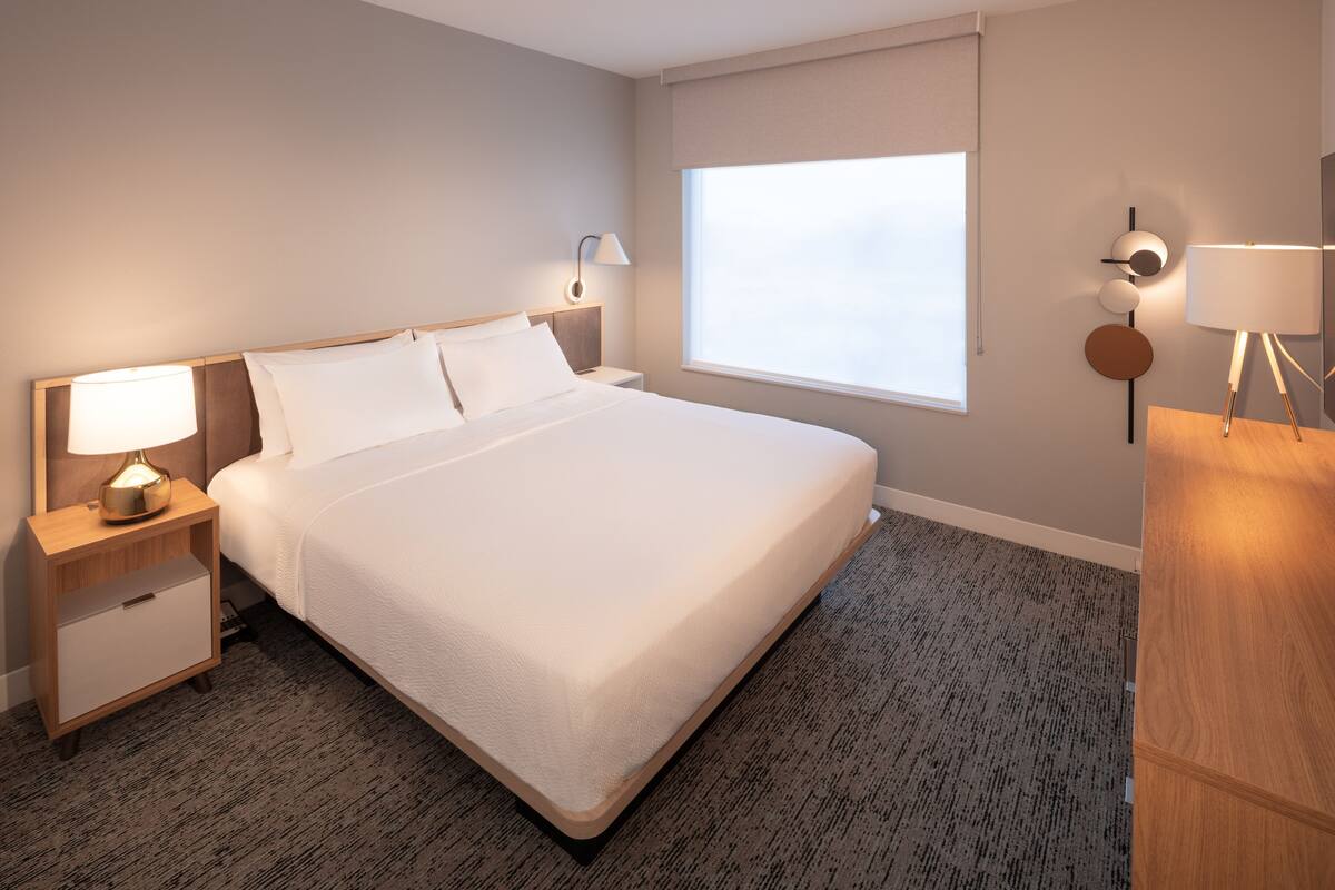 Habitación de hotel con una cama grande, mesitas de noche con lámparas, un ventanal con vistas y suelo alfombrado.