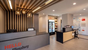 A hotel reception area with a modern design, featuring a reception desk labeled "HELLO Ellensburg," a refrigerator stocked with beverages, and a wall displaying a map and welcome sign.