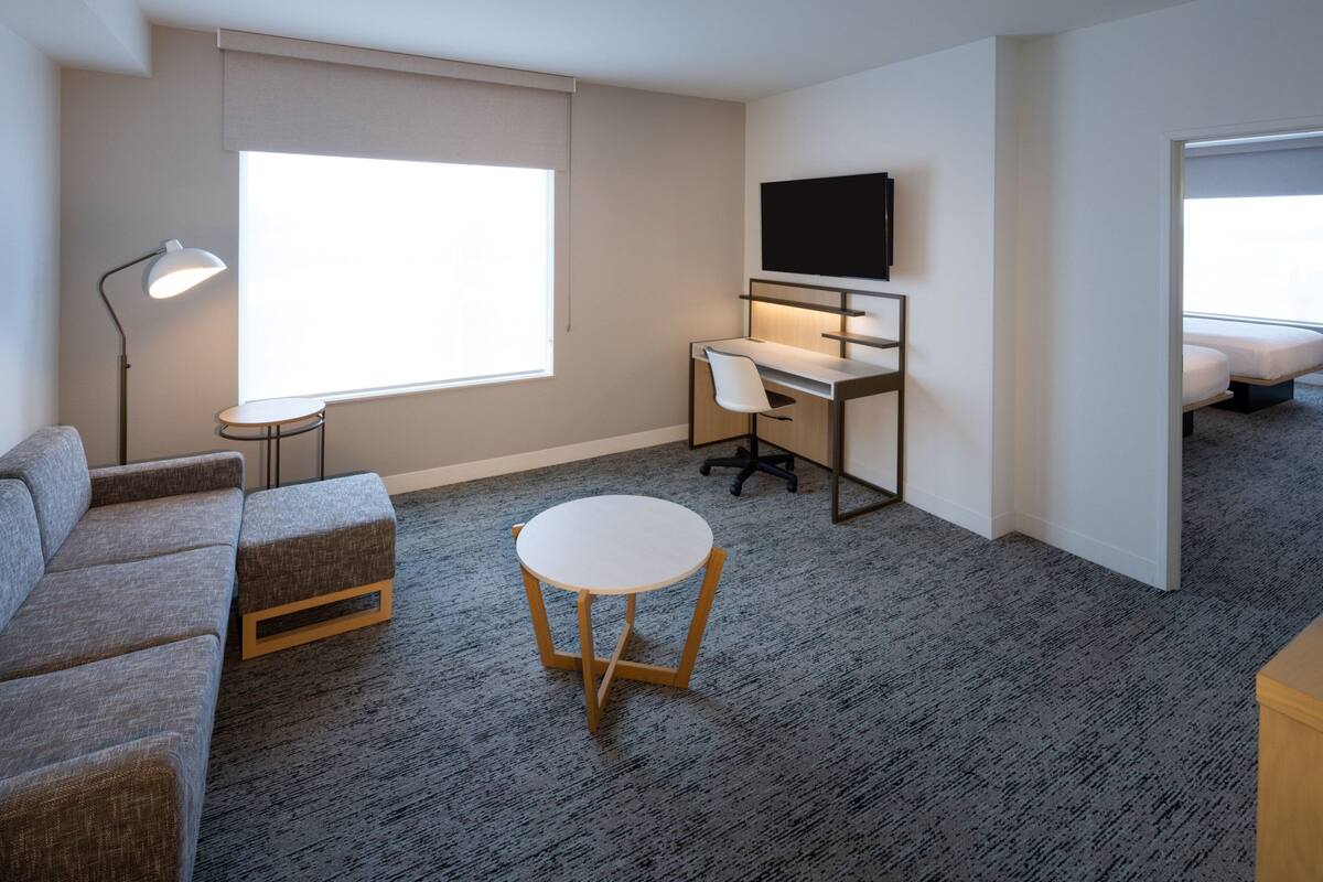 Una habitación de hotel moderna con un sofá gris, una pequeña mesa redonda, un escritorio con una silla, un televisor montado en la pared y un amplio ventanal que proporciona luz natural.
