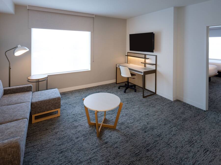 Una habitación de hotel moderna con un sofá gris, una pequeña mesa redonda, un escritorio con una silla, un televisor montado en la pared y un amplio ventanal que proporciona luz natural.