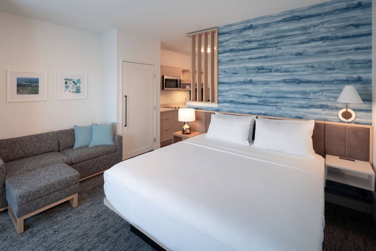 Habitación de hotel con una cama grande, un sofá, una mesita, una lámpara y una pared con un diseño abstracto azul.
