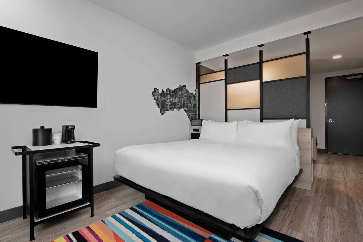 Una habitación de hotel moderna con una cama grande, un televisor de pantalla plana en la pared, una cafetera y un refrigerador pequeño.