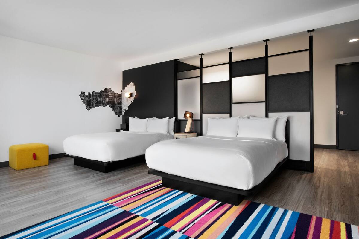 Una habitación de hotel moderna con dos camas, un colorido tapete a rayas, una original obra de arte en la pared y un pequeño otoman amarillo.