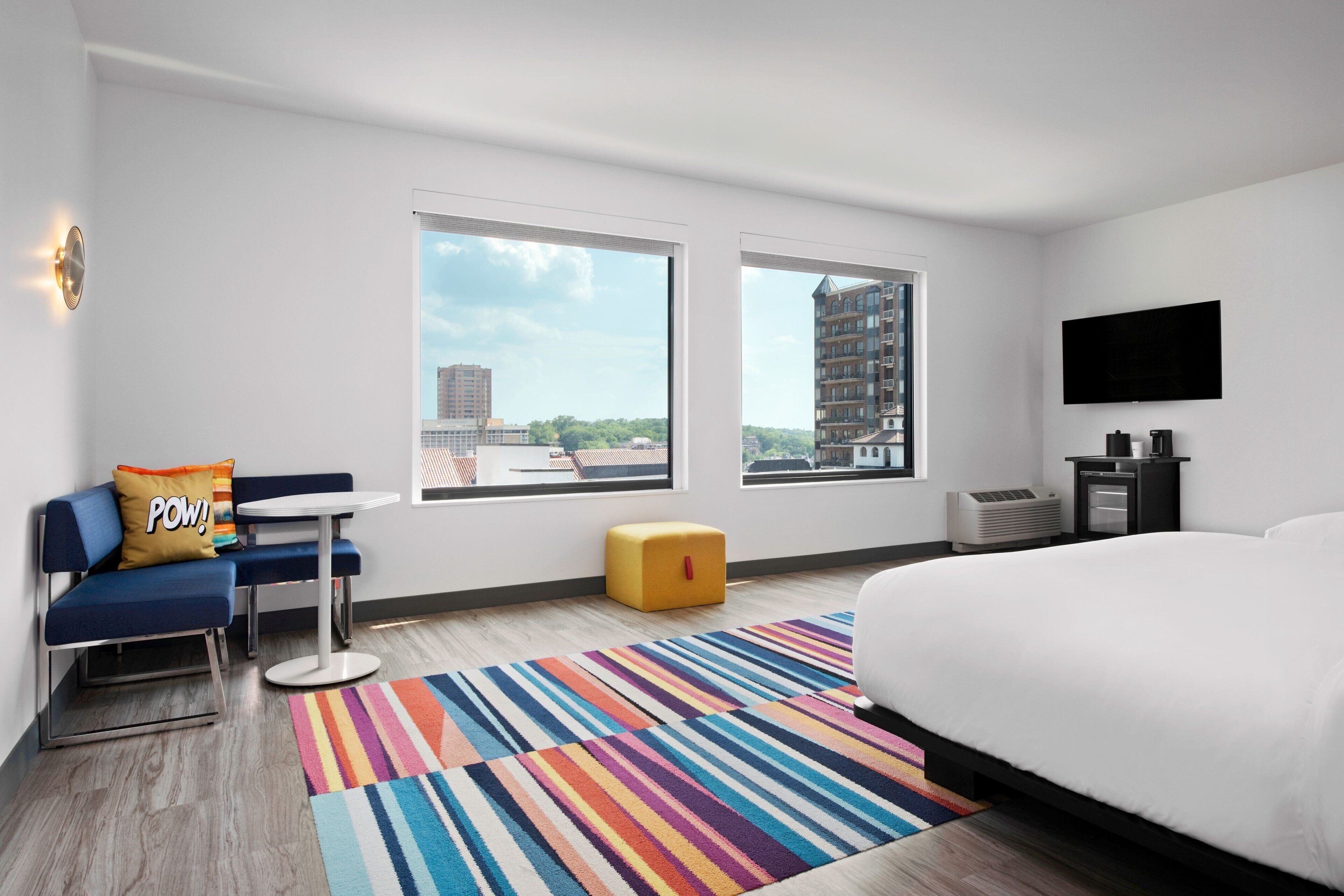 A modern hotel room with a bed, a sofa, a small table, and two windows offering city views.