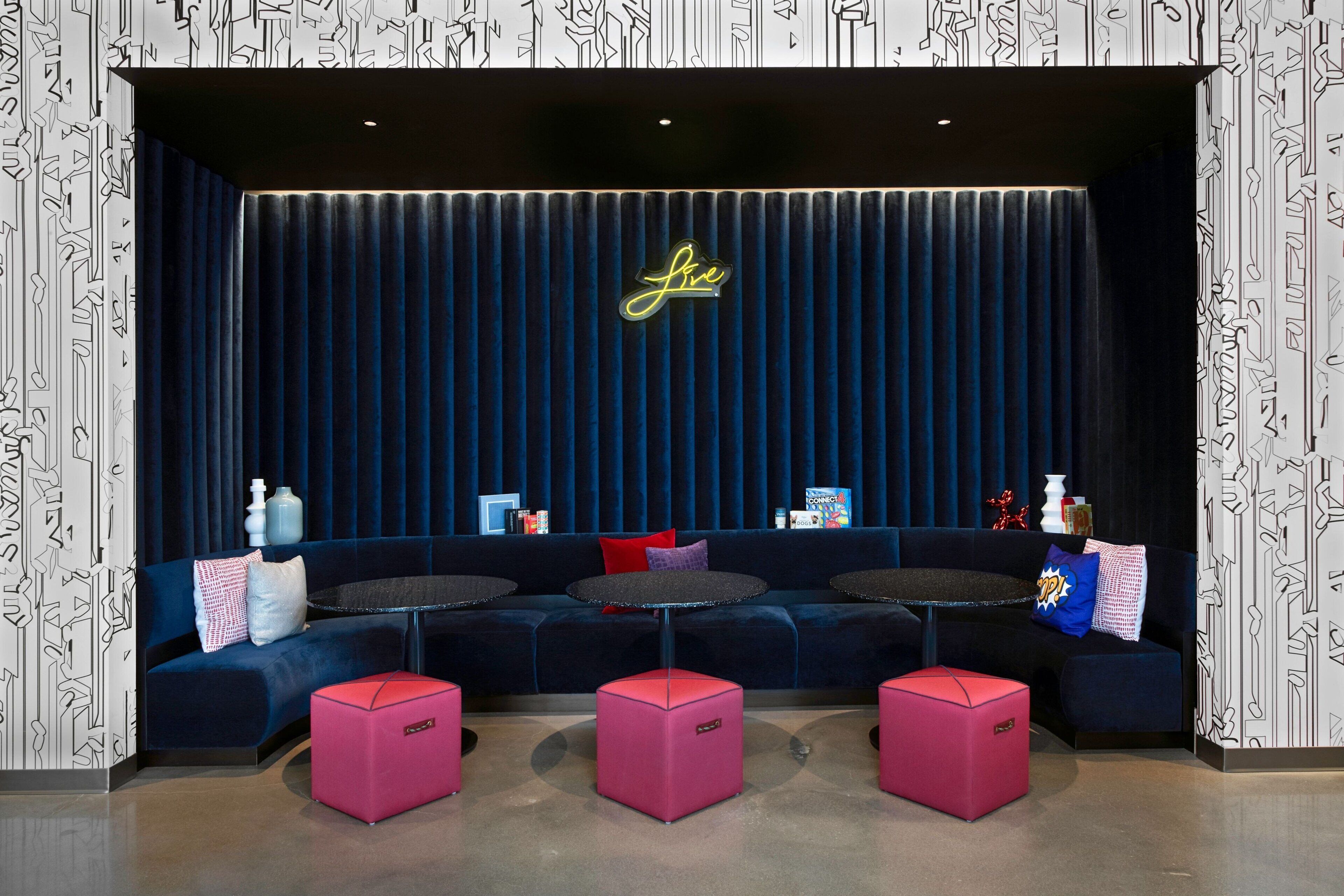 A modern lounge area with a dark blue sofa, pink ottomans, and a neon sign.