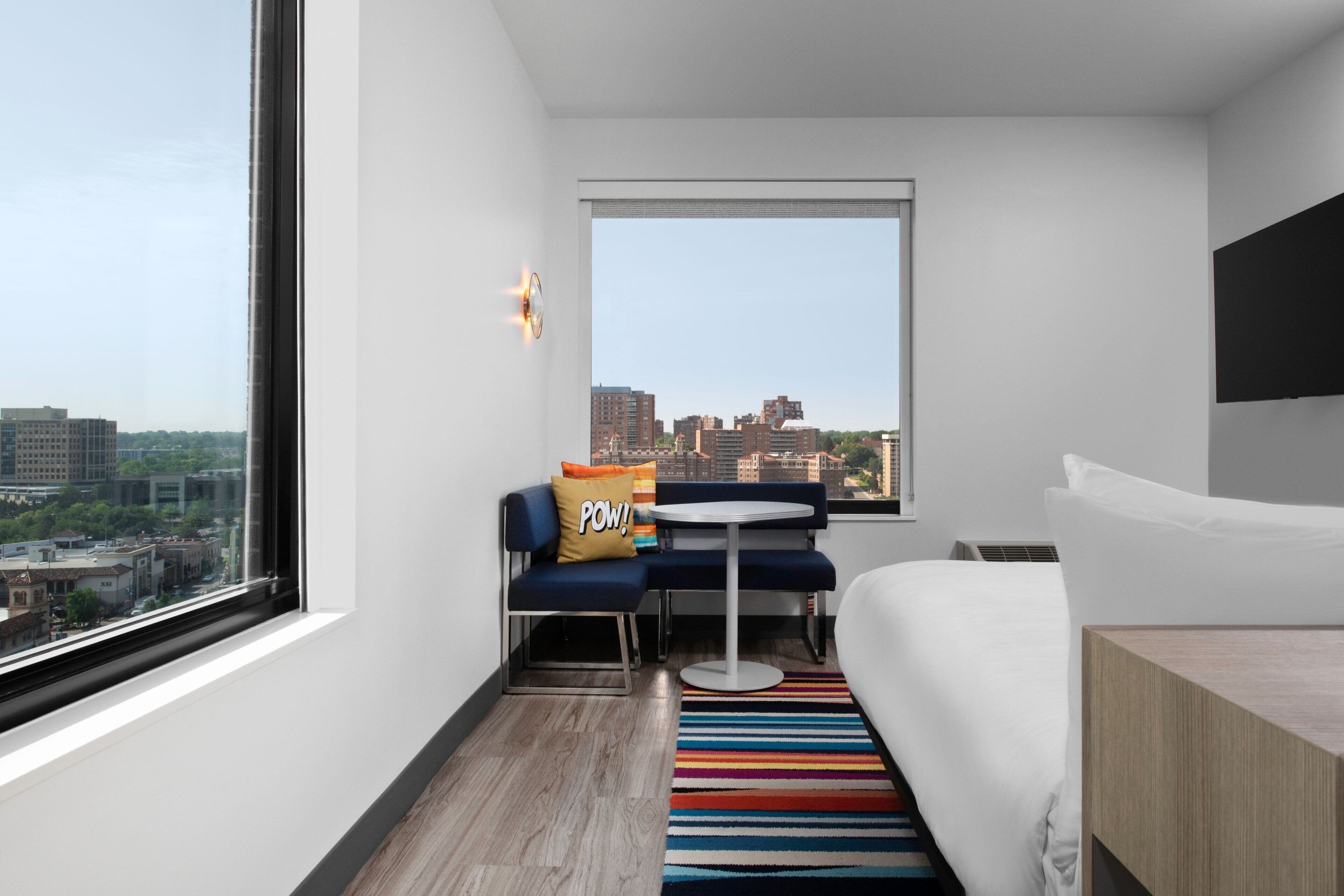 A modern hotel room with a large window, a bed, a small round table, and a sofa with a city view.
