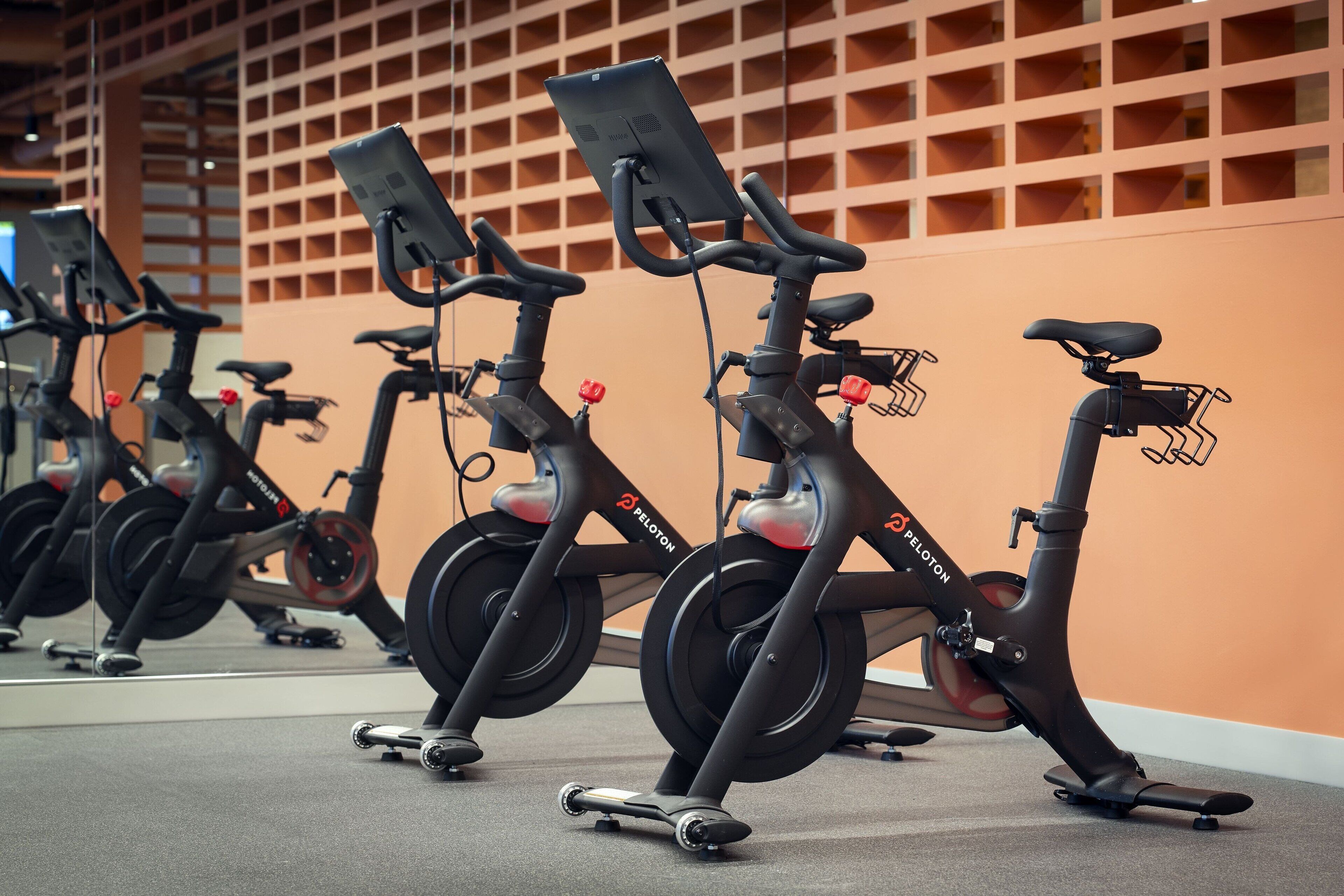 A row of stationary bikes in a gym with a mirrored wall and a grid-patterned ceiling.