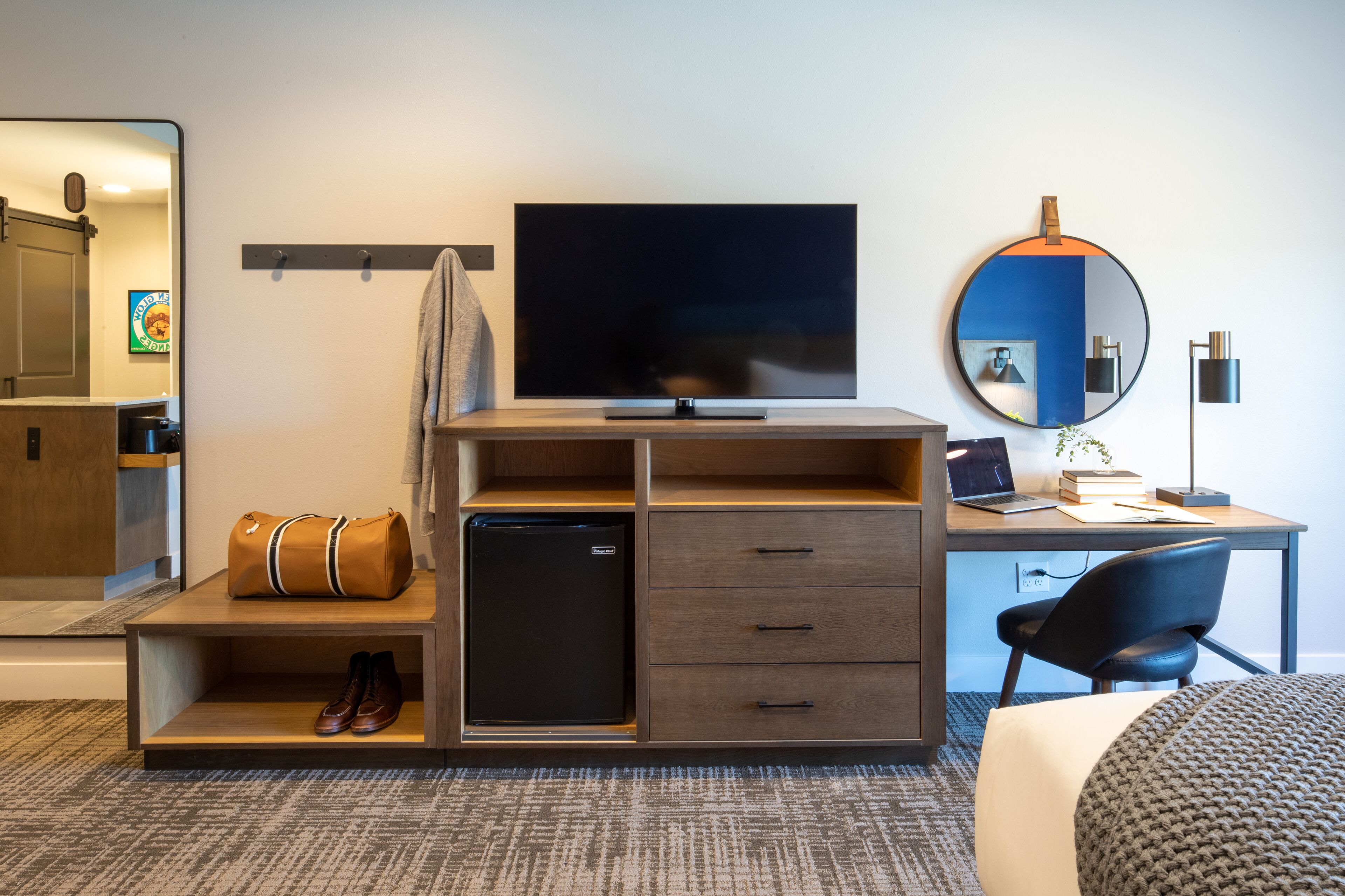 A hotel room with a TV, a desk with a laptop, a small refrigerator, a mirror, and a chair.