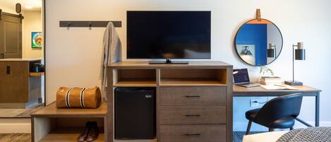 A hotel room with a TV, a desk with a laptop, a small refrigerator, a mirror, and a chair.
