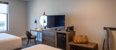 Habitación de hotel con una cama, un escritorio con computadora, televisión, cafetera y un bolso sobre una mesita de noche.