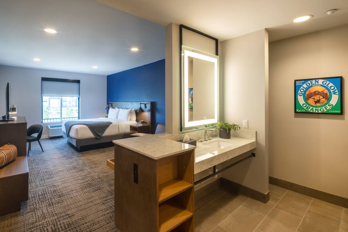 Ein modernes Hotelzimmer mit einem großen Bett, einer Sitzecke mit einem Sessel, einem Schreibtisch, einem Schminktisch mit Spiegel und einem Badezimmer mit Waschbecken und Toilette.