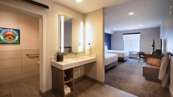 Un baño de hotel con un espejo grande, lavamanos y un perchero. En la habitación contigua hay una cama y un escritorio.