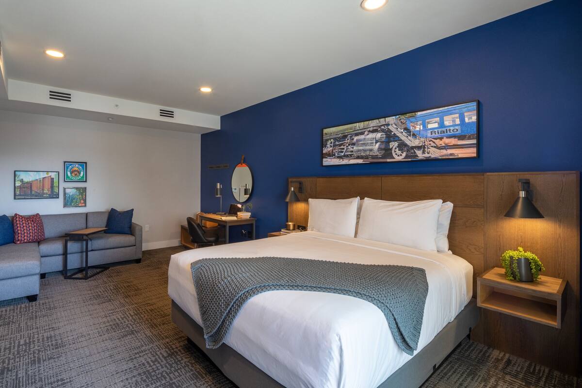 Ein Hotelzimmer mit einem großen Bett, einer Couch, einem Schreibtisch und einem kleinen Tisch. An der Wand hängt ein Gemälde und auf dem Nachttisch steht eine Pflanze.