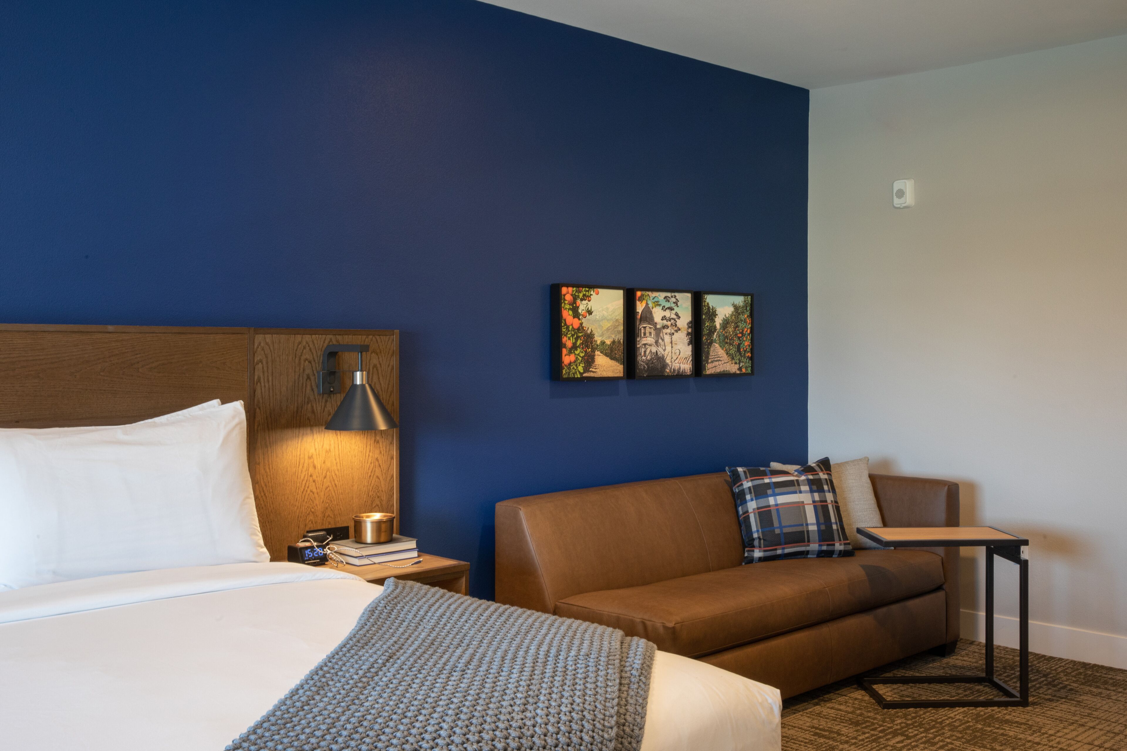 Habitación de hotel con cama, sofá de cuero marrón, una mesita y una obra de arte colgada en la pared.