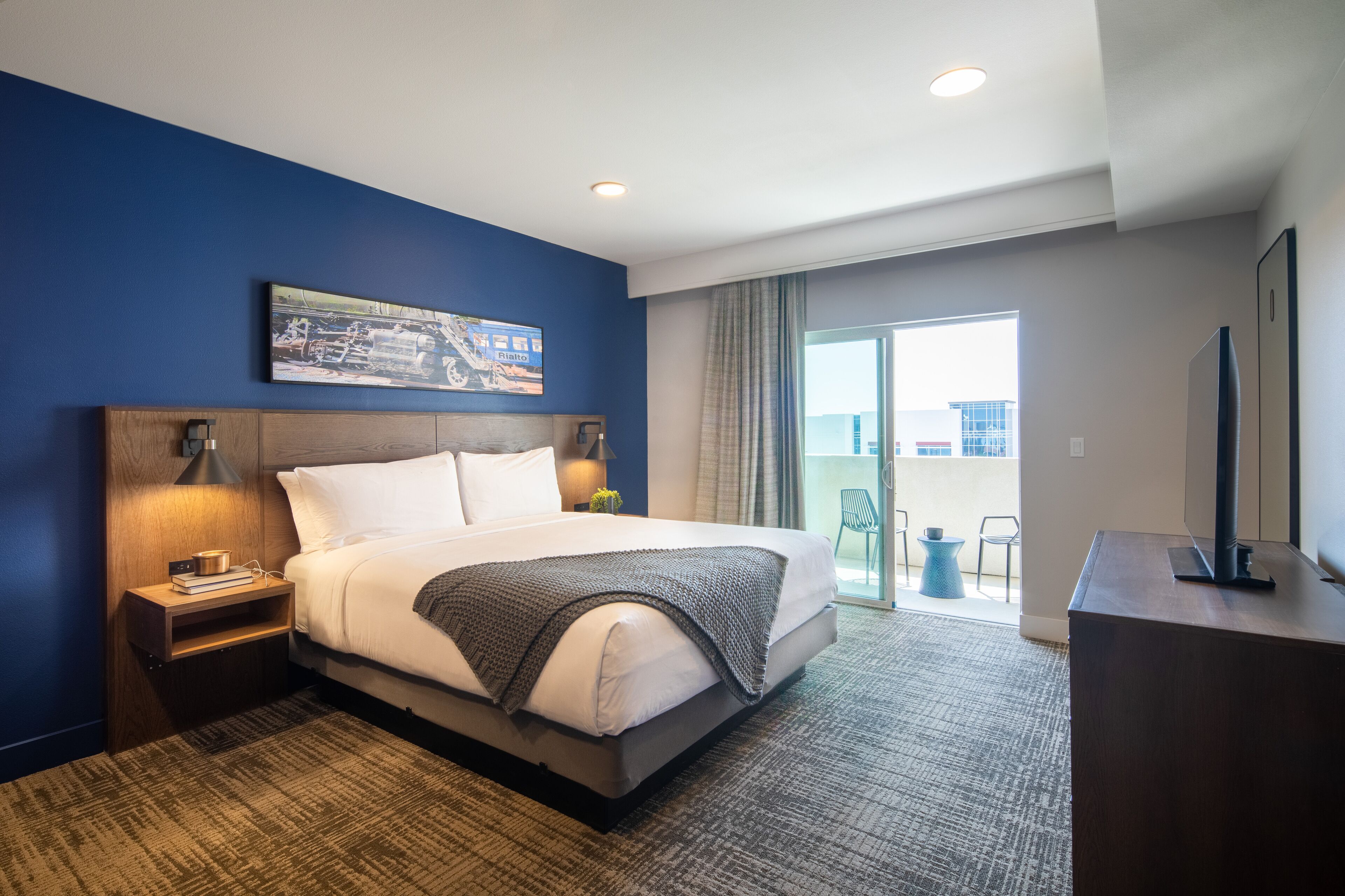 A hotel room with a large bed, a nightstand, a desk, and a view of the outside balcony.