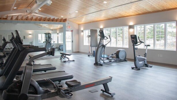 Ein Fitnessstudio mit Trainingsgeräten, großen Fenstern und einer Holzbalkendecke.