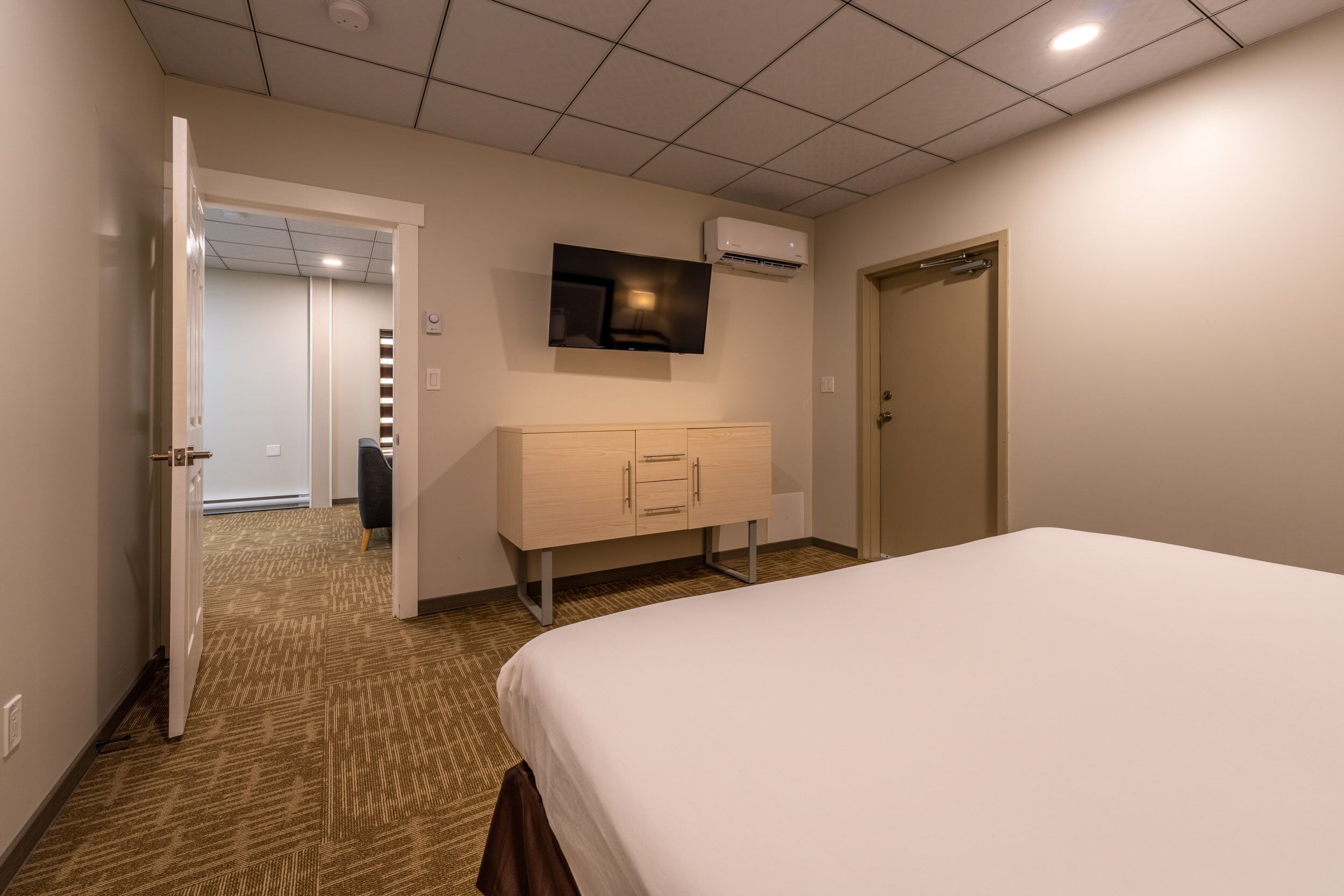 A hotel room with a bed, a TV mounted on the wall, a wooden cabinet, and an air conditioning unit.