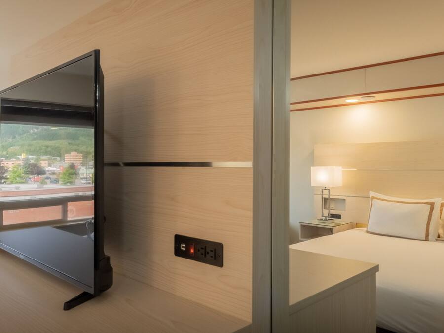 Una habitación de hotel con cama, mesita de noche, lámpara y televisión montada en la pared.