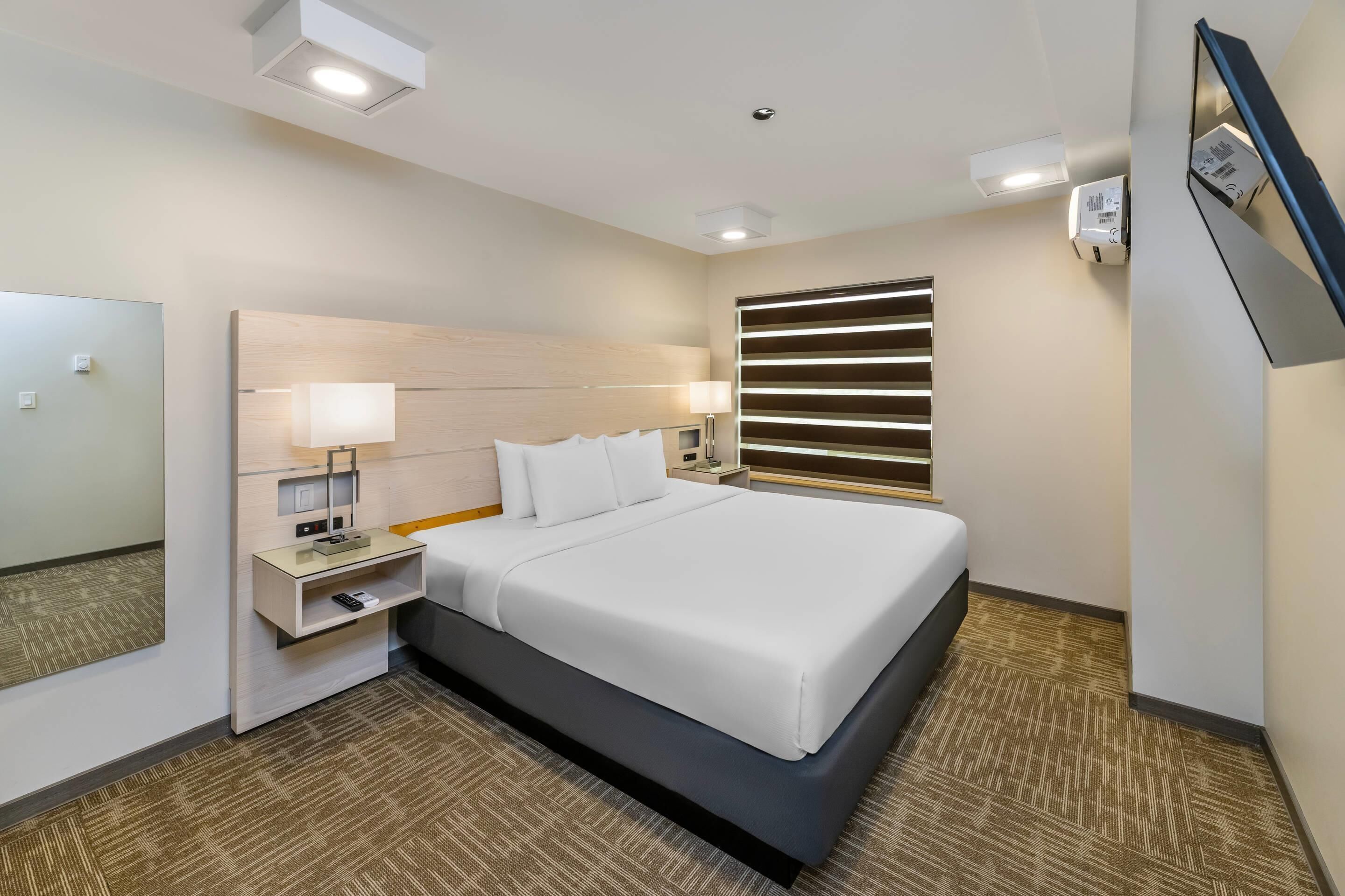 Una habitación de hotel con una cama grande, mesitas de noche, un espejo y un televisor montado en la pared.
