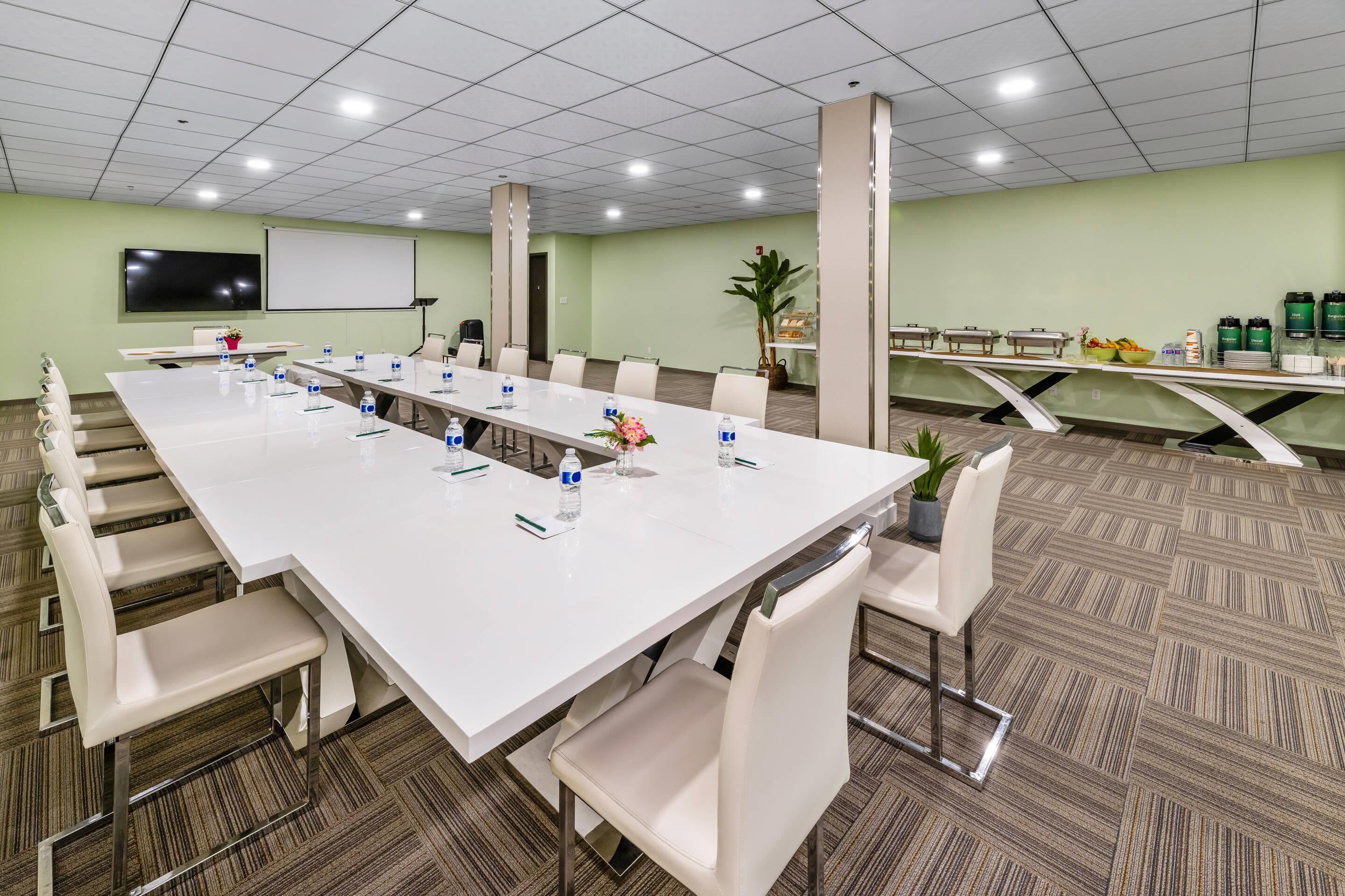 Een vergaderruimte met een lange tafel, stoelen, een scherm voor een beamer en een buffetgedeelte met eten en drinken.