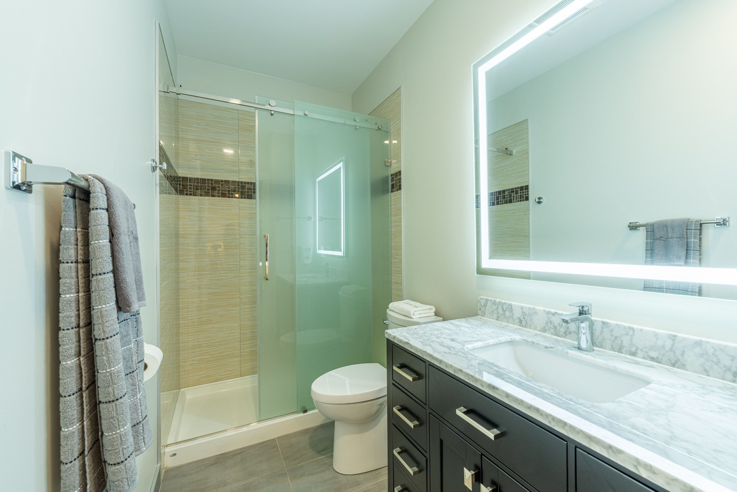 Une salle de bain moderne avec une douche à parois de verre, des toilettes blanches, un lavabo en marbre et un grand miroir.