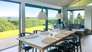 A dining area with a wooden table, black chairs, and woven placemats. There is a sofa with striped cushions and a view of a green landscape through large windows. - 3 bedroom stunning home in Haderslev (Haderslev)
