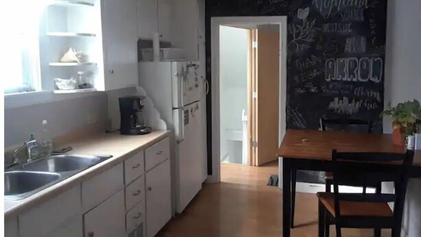 Une cuisine moderne avec des meubles blancs, une machine à café et un mur noir où l’on peut écrire à la craie.